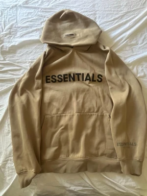 Beige Essentials hoodie med tryck i storlek M - Säljer en beige hoodie från Essentials med svart tryck på bröstet och liten logga på ärmen. Hoodien har stor huva och magficka framtill. Tillverkad i mjuk bomullsblandning, perfekt för en avslappnad streetwear-look.
