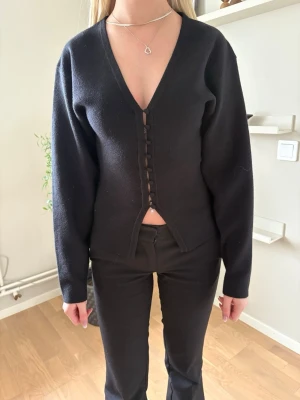 Svart kofta med knappar och v-ringning - Stilren svart kofta med djup v-ringning och dekorativa klädda knappar framtill. Långärmad modell i mjukt material som sitter snyggt på kroppen. Perfekt att matcha med både jeans och kjol för en trendig look.