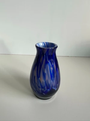 Blå mönstrad glasvas - Säljer en unik vas i blått glas med virvlande mönster i olika blå nyanser. Vasen har en mjukt rundad form och smal öppning, perfekt som dekoration eller för blommor. Materialet är glas och färgen skiftar mellan mörkblått och ljusare vita och rosa toner. Höjd 12cm, diameter 6cm. Mycket fint skick!