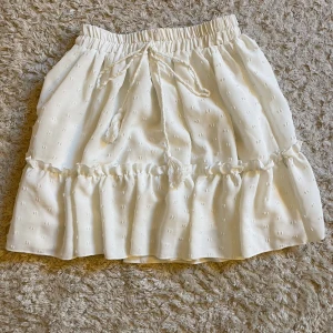 Vit volangkjol med snörning - Söt vit kjol med volang och snörning i midjan. Kjolen har en lätt struktur i tyget och resår i midjan för skön passform. Perfekt för en fräsch och somrig look.