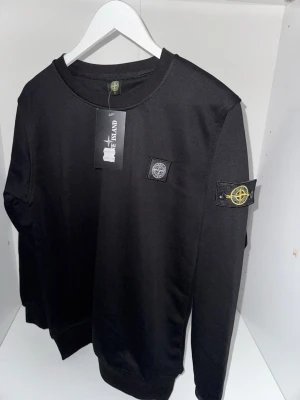 Svart sweatshirt från Stone Island - Snygg svart sweatshirt från Stone Island med klassisk rund hals och ikoniska logopatchen på vänster ärm. Tröjan har ribbade muddar vid ärmslut och nederkant samt en diskret logga på bröstet. Perfekt för en stilren och trendig look.  Det står på tröjan att det är storlek M men den passar som S ( Den passar i storlek S och M)