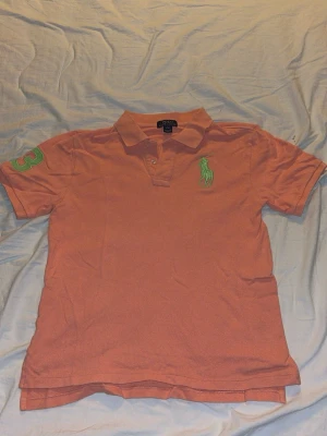 Korallfärgad pikétröja Ralph Lauren - Säljer en korallfärgad pikétröja från Ralph Lauren med klassisk krage och korta ärmar. Tröjan har en grön broderad logga på bröstet och en grön siffra 3 på ärmen. Perfekt till en avslappnad och stilren look.