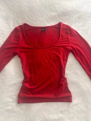 Röd långärmad topp från Gina Tricot - En enkel och snygg röd långärmad topp från Monki med rundad, djup urringning. Tillverkad i mjuk bomull som sitter skönt mot kroppen. Perfekt att styla med jeans eller kjol för en clean look.