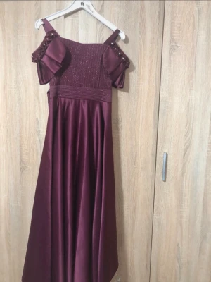 Vinröd långklänning med pärldetaljer - Vinröd långklänning med axelbandsdesign och dekorativa pärlor på ärmarna. Klänningen har en smockad överdel och faller snyggt i ett glansigt tyg. Perfekt för dig som vill sticka ut med eleganta detaljer och en klassisk siluett. Använd 2-3 gånger, klänningen är mer till bröllop eller fester, jag köpte den utomlands. Jag kan skicka bild när jag hade på mig den samt storleken. Pris går att diskuteras:)