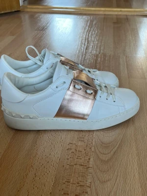 Vita Valentino sneakers med roségulddetalj - Snygga vita sneakers från Valentino med bred roséguldfärgad metallicdetalj över snörningen. Skorna är låga och har klassisk siluett med vita skosnören och diskret logga på plösen. Tillverkade i läder för en lyxig känsla och stilren look. Dessvärre har de någon skråma (se bild) Skorna ser mer roséfärgade ut i ljuset och guldigare när det är mörkt