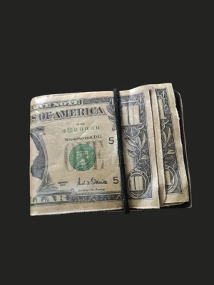 Plånbok  - Unik plånbok med tryck av amerikanska dollarsedlar. Plånboken har en klassisk rektangulär form och hålls ihop med ett svart resårband. Materialet ser ut som konstläder eller plast med realistiskt sedelmotiv. Perfekt för dig som gillar statement-accessoarer.