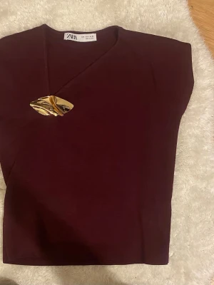 Vinröd topp från Zara med gulddetalj - Säljer en vinröd topp från Zara med korta ärmar och en unik guldfärgad metall-detalj vid halsringningen. Toppen har en snygg, figurnära passform och V-ringning. 