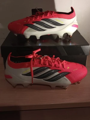 Adidas Predator elite fotbollsskor HELT NYA!!! - Säljer ett par Adidas Predator elite FG fotbollsskor i en snygg kombination av röd, vitt och svart. Skorna har klassiska tre ränder på sidan, snörning och dobbar i metall för bästa grepp på planen. Ovandelen är i syntetmaterial och designen är riktigt modern och sportig. 