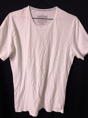 Vit t-shirt från Calvin Klein - Klassisk vit t-shirt från Calvin Klein med rund halsringning och diskret logga på insidan av kragen. Tillverkad i mjuk bomull och har en enkel, clean design som passar till allt. Perfekt basplagg för dig som gillar stilrena och tidlösa looks.
