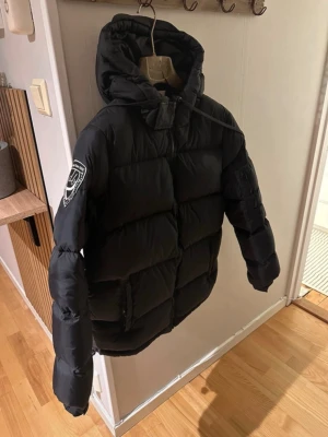 Svart dunjacka med huva - Säljer en svart pufferjacka med stor huva och patch på ena ärmen. Jackan har dragkedja framtill, två stora fickor och quiltad design. Perfekt för kalla dagar och riktigt skön att bära. Passar dig som vill ha en stilren och varm vinterjacka.