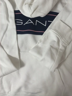 Vit hoodie från GANT med logga - Snygg vit hoodie från GANT med stor blå logga och rosa detalj på bröstet. Klassisk huva och magficka framtill. Tillverkad i mjukt bomullsmaterial som känns skönt mot huden. Perfekt för en avslappnad och trendig look.