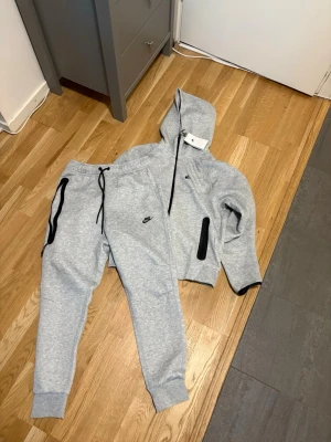 Nike tech S - Snygga gråa mjukisbyxor och hoodie från Nike med svarta detaljer och dragsko i midjan. Byxorna har ribbade muddar vid bensluten och praktiska fickor med dragkedja. Perfekta för chill eller träning, matchar med hoodie för en clean look. Helt oanvända helt nya aldrig tagit på mig dom, lite för liten för mig men annars ser allt väldigt bra ut.