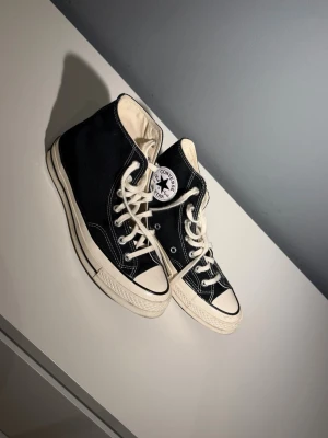 Svarta Converse Chuck Taylor High - Klassiska svarta Converse Chuck Taylor All Star high tops med vit sula och tå, vita snören och kontrasterande sömmar. Ikonisk logga på sidan och canvasmaterial som ger en tidlös och cool look. Perfekta för dig som gillar streetstyle och retrovibbar.