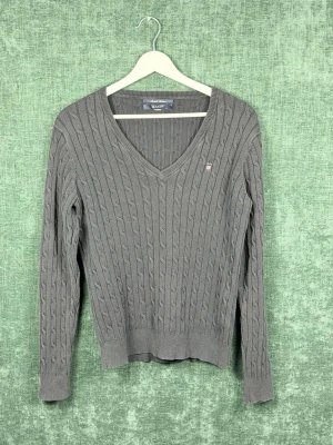 Grå kabelstickad tröja från GANT - Snygg grå kabelstickad tröja från GANT med V-ringning och diskret logga på bröstet. Tröjan har långa ärmar och ribbade muddar vid ärmslut och nederkant. Perfekt till jeans eller kjol för en klassisk look.