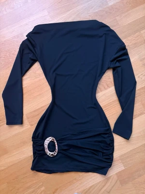 Svart oneshoulder kortklänning - Snygg svart kortklänning med offshoulder-design och långa ärmar. Klänningen har en dekorativ oval detalj i midjan och figurnära passform. Jag har endast provat klänningen men tyckte inte den passade min stil. Skriv om ni vill ha fler bilder! 