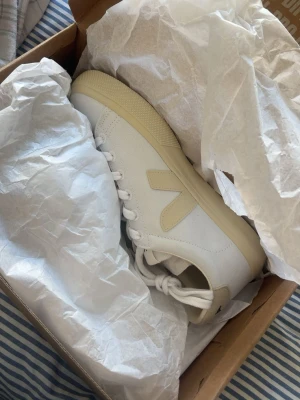 Vita Veja sneakers med beige detaljer - Snygga vita Veja sneakers med beige detaljer och klassisk V-logga på sidan. Skorna har lågt skaft, vita snören och beige sula. Tillverkade i canvas och passar perfekt till en clean och stilren look.
