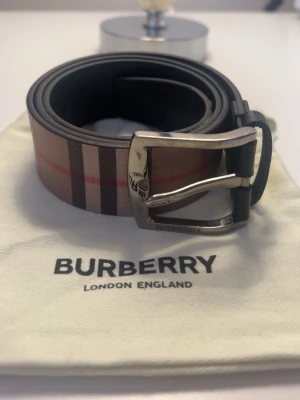 Burberry bälte i skinn - Snyggt bälte från Burberry med klassiskt randigt mönster i beige, svart, rött och vitt. Bältet har en silverfärgad metallspänne och är gjort i skinn. Köpt av Melker Ahlquist 2023/24 via Ig dm. 