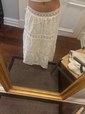 Vit långkjol med spetsdetaljer - Säljer en superfin vit långkjol med broderade spetsdetaljer och knytband i midjan. Kjolen har romantiska volanger och ett luftigt, bohemiskt uttryck. Perfekt för varma dagar och sommarens alla äventyr.