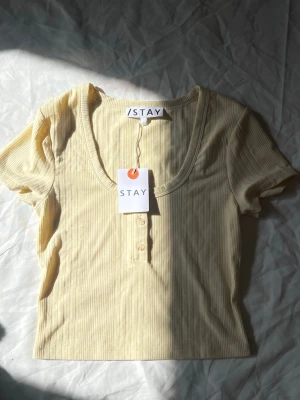Ljusgul ribbad topp från Stay - Kortärmad topp från Stay i ljusgul ribbad bomull med rundad v-ringning och knappar framtill. Snygg croppad passform och diskreta ränder i materialet. Perfekt till jeans eller kjol för en fräsch look.