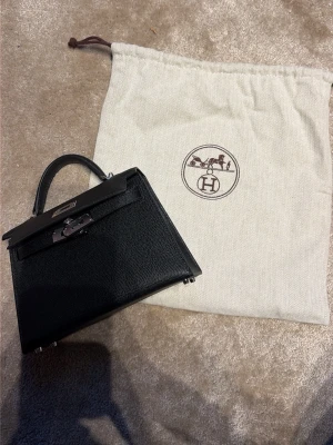 Svart Kelly Mini handväska - Säljer en svart  Kelly Mini handväska i exklusivt läder med silverfärgade metalldetaljer och klassiskt vridlås. Väskan har ett elegant topphandtag och kommer med original dustbag. Perfekt för dig som vill ha en ikonisk och lyxig accessoar.