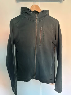 Mörkgrön hoodie från Sail Racing - Mörkgrön hoodie från Sail Racing med dragkedja framtill och en bröstficka med zip. Tillverkad i mjukt material och har en klassisk huva. Perfekt för en avslappnad och sportig stil. Hör av er vid frågor eller lägg ett prisförslag!