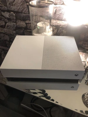 Xbox One S vit spelkonsol - Säljer en Xbox One S spelkonsol i snygg vit färg med stilren, rektangulär design och ventilerad ovansida. Perfekt för gaming och streaming. Konsolen är tillverkad i plast och har klassisk Xbox-logga på framsidan.