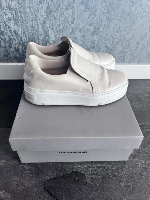 Vita slip-on sneakers från Vagabond - Stilrena vita slip-on sneakers från Vagabond med chunky sula och minimalistisk design. Skorna är tillverkade i slätt läder och har en diskret logga på hälen. Perfekta för dig som gillar enkel och clean stil.