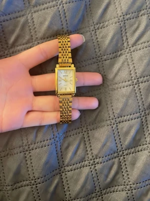 Guldfärgad fyrkantig klocka Seiko - Stilren guldfärgad klocka från Seiko med fyrkantig urtavla och länkarmband. Urtavlan är vit med romerska siffror och tunna visare. Klassisk och elegant design som passar till många olika stilar.