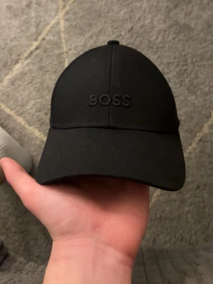 Svart keps från BOSS - Snygg svart keps från BOSS med broderad logga framtill. Klassisk böjd skärm och justerbar rem bak för perfekt passform. Tillverkad i ett mjukt och slitstarkt material som känns bekvämt hela dagen.