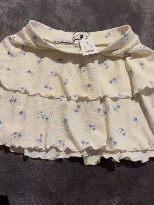 Vit blommig volangkjol  - Supersöt kort kjol från H&M i vit med små blå och gula blommor. Kjolen har volangdetaljer och resår i midjan för en bekväm passform. Perfekt för sommarens alla tillfällen!