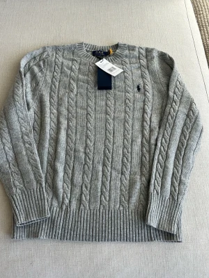 Grå kabelstickad tröja Polo Ralph Lauren - Snygg grå kabelstickad tröja från Polo Ralph Lauren med rund halsringning och ribbade muddar. Tröjan har det klassiska broderade logotypen på bröstet och är tillverkad i mjukt garn. Perfekt för en stilren och avslappnad look.