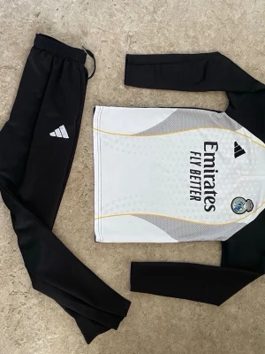Real Madrid träningsset Adidas svart/vit - Snyggt träningsset från Adidas med Real Madrid-tryck. Tröjan är vit med svarta ärmar, gula detaljer och klubbmärke, samt texten 'Emirates Fly Better' på bröstet. Byxorna är svarta med Adidas-logga. Perfekt för fotbollsträning eller gymmet.