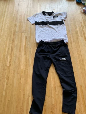 The North Face svartvitt set - Snyggt set från The North Face med en kortärmad vit tröja med svart rand och knappar vid halsen samt matchande svarta byxor med resår i midjan. Båda plaggen har tydliga logotyper tryckta framtill. Perfekt för en avslappnad och sportig stil.