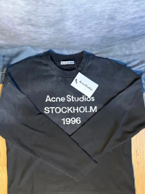 Svart sweatshirt Acne Studios - Svart långärmad sweatshirt från Acne Studios med vit text 'Acne Studios STOCKHOLM 1996' på bröstet. Klassisk rund halsringning och avslappnad passform. Perfekt för en trendig streetwear-look.
