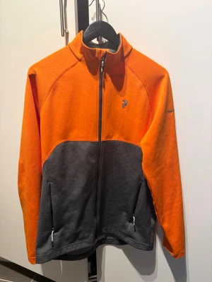 Orange och grå ziptröja Peak Performance - Snygg ziptröja från Peak Performance i orange och mörkgrått. Tröjan har hög krage, hel dragkedja framtill och två fickor med dragkedja. Perfekt för lager-på-lager och outdoor. Broderad logga på bröstet och ärmen.