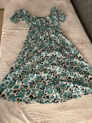 Blommig långklänning med puffärm - Långklänning med grönt och blått blommönster på vit botten. Klänningen har puffiga kortärmar med resår och en smickrande, något figurnära passform. Perfekt för sommarens alla tillfällen!