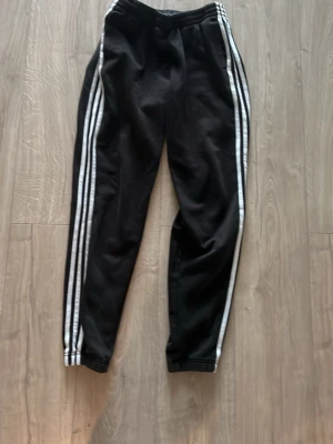 Svarta Adidas mjukisbyxor med ränder - Säljer ett par klassiska svarta mjukisbyxor från Adidas med vita ikoniska tre ränder längs benen. Byxorna har resår i midjan och vid bensluten, samt en avslappnad passform. Perfekta för chill eller träning.
