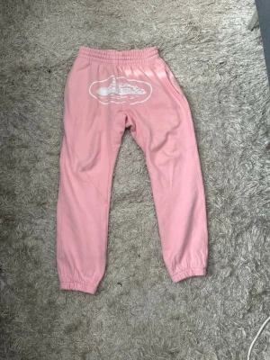 Rosa corteiz mjukisbyxor - Säljer ett par ljusrosa corteiz mjukisbyxor med resår i midjan och benslut. Byxorna har ett vitt tryck framtill och sidofickor samt en bakficka. Perfekta för chill dagar och riktigt sköna att ha på sig. Materialet känns mjukt och bekvämt. Passar perfket till den matchande hoodien. Hela settet/tracksuiten finns i min profil som en egen annons.