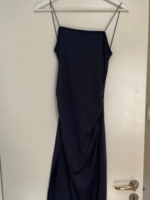 Marinblå långklänning med öppen rygg - Snygg marinblå långklänning med tunna axelband och draperad detalj framtill. Klänningen har en öppen rygg med twistad knutdetalj och är tillverkad i ett mjukt, följsamt material som ger en elegant siluett.