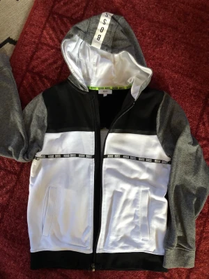 Hugo Boss hoodie med dragkedja - Snygg hoodie från Hugo Boss i svart, vitt och grått. Hoodien har dragkedja framtill, två fickor och en huva med BOSS-tryck. Detaljer med Hugo Boss-logga på bröstet och längs med bröstet. Perfekt för en avslappnad och trendig stil.passar till 12 år. Använd 1gång