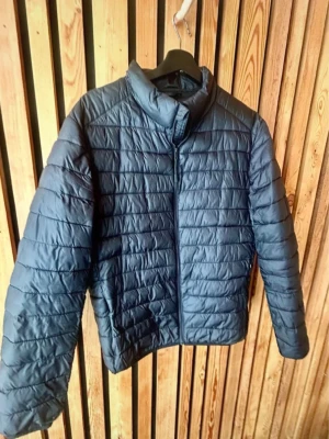 Svart Dunjacka från Zara - Snygg mörkblå pufferjacka från Zara med quiltad design och hög krage. Jackan har dragkedja framtill och är tillverkad i lätt, vadderat material som håller värmen. Perfekt för kalla dagar och enkel att matcha med olika outfits.