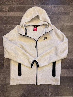 Nike tech fleece - Snygg ljusgrå hoodie från Nike med dragkedja och svarta detaljer längs dragkedjan och fickorna. Klassisk Nike-logga på bröstet. Utrustad med huva och två fickor framtill. Perfekt för en sportig och avslappnad stil.