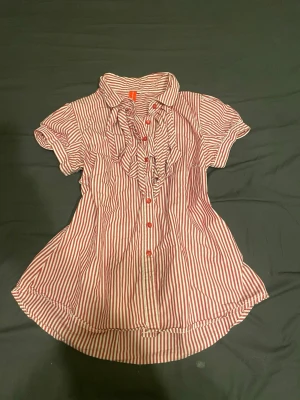 Rödvit randig blus med volang - Snygg blus från H&M med röda och vita ränder, volangdetaljer längs knäppningen och röda knappar. Kortärmad modell med klassisk krage och lätt utsvängd passform. Perfekt för en trendig och avslappnad look. Passar fint på mig som är xs/s