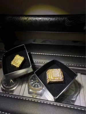 2 st Guld fyrkantig ring med stenar - Säljer 2 st riktigt fet guldfärgade ringar med fyrkantig form, täckt av gnistrande stenar i flera lager. Ringarna har en bred design och ser exklusiva ut, perfekt för dig som vill sticka ut. Levereras i snygg ask.