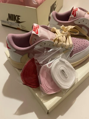 Nike Air Jordan 1 Low rosa/vit - Nike Air Jordan 1 Low sneakers i rosa och vitt med beige detaljer och röd Nike-logga på plösen. Ovandel i mocka och läder, perforerad tå och klassisk swoosh i ljusgul. Skorna kommer med extra skosnören i rött, rosa och vitt. Perfekt för dig som gillar unik stil.