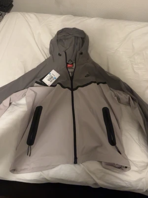 Nike grå vindjacka med huva - Snygg grå vindjacka från Nike med svart dragkedja och detaljer. Jackan har huva, två stora fickor med svarta dragkedjor och en sportig look. Tillverkad i lätt och tåligt material, perfekt för blåsiga dagar. Nike-logga på bröstet och modern passform.