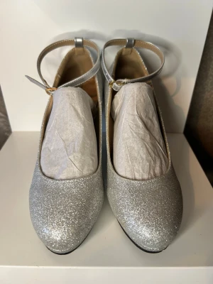 Silvriga glittriga klackskor med rem - Silvriga klackskor med glitter och rund tå. Skorna har en smal ankelrem med spänne och en medelhög klack. Perfekta för dig som vill sticka ut och addera lite extra bling till din outfit. 