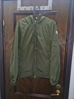 Olivgrön vindjacka från Moncler - Snygg olivgrön vindjacka från Moncler med huva och dragkedja framtill. Jackan har två sidofickor med tryckknappar, elastiska muddar vid ärmsluten och Moncler-logga på ärmen. Tillverkad i lätt, vindtätt material som passar perfekt för blåsiga dagar.