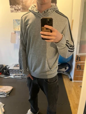Grå hoodie från Adidas med svarta ränder - Säljer en grå hoodie från Adidas med klassiska svarta ränder längs ärmarna och Adidas-logga på bröstet. Hoodien har huva och en stor magficka. Tillverkad i mjukt bomullsmaterial, perfekt för en avslappnad streetwear-look.