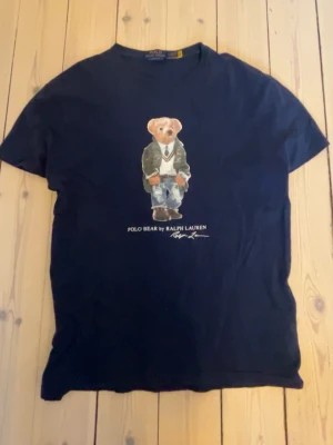 Navy blå Polo Bear t-shirt från Ralph Lauren - Klassisk navy t-shirt från Ralph Lauren med Polo Bear-tryck på bröstet. T-shirten har rund hals, korta ärmar och är gjord i mjuk bomull. Perfekt för dig som gillar stilrena plagg med en unikt tryck.